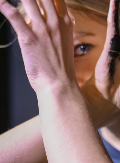 Hidden Polution (photograph of a girl looking between her hands towards the camera, with black paint on one of her hands)