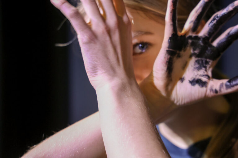 Hidden Polution (photograph of a girl looking between her hands towards the camera, with black paint on one of her hands)